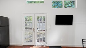 A room with a black refrigerator, a white door with glass panels, a black mat at the door, a flat-screen TV mounted on the wall, and a small window above the door. - Austin Cozy Tiny Home | Fire Pit | Pet-Friendly (Austin)