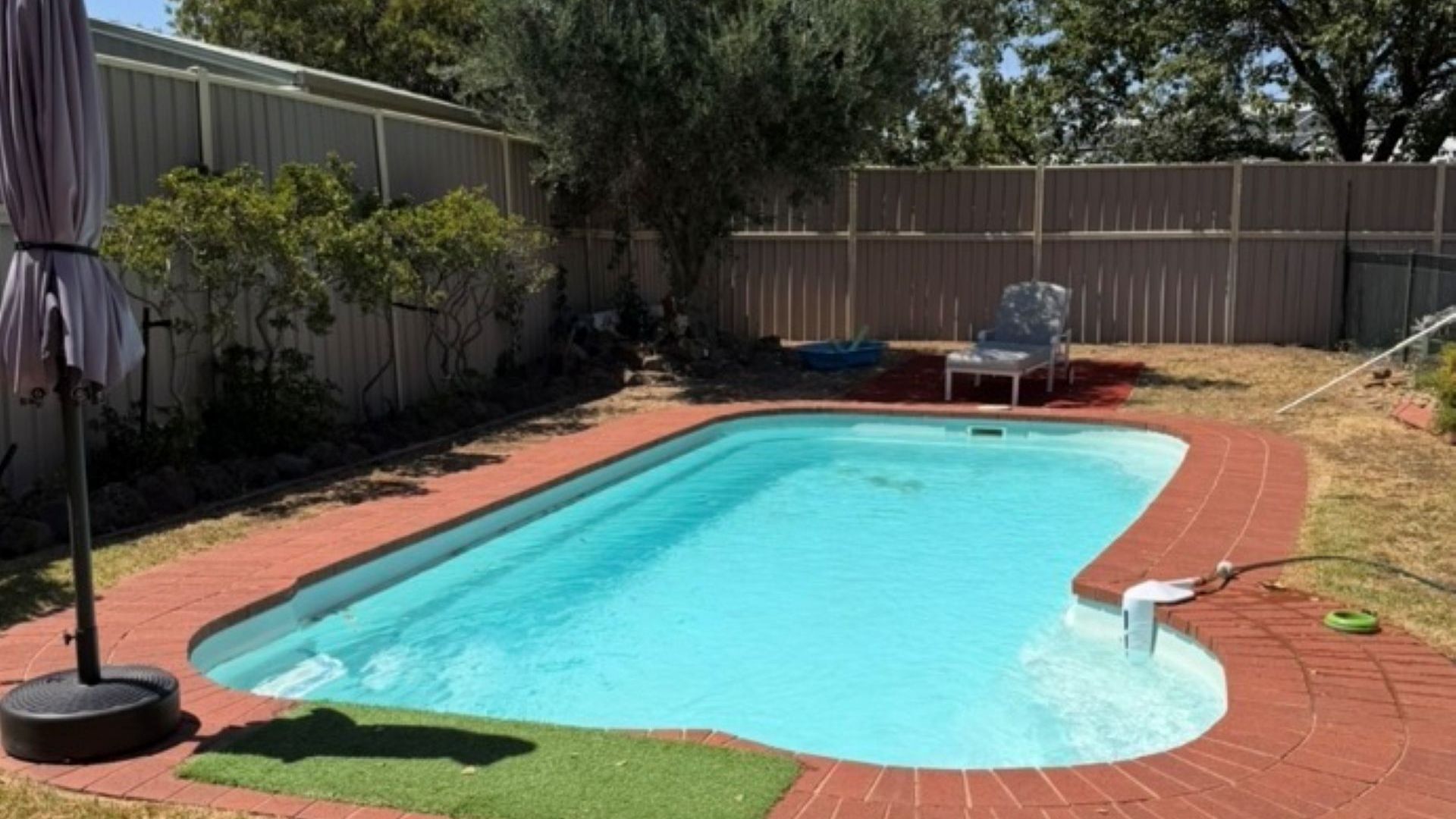 Ein rechteckiger Pool mit Ziegelkanten, umgeben von einer Rasenfläche und einem Holzzaun.