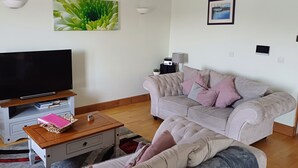 Uma sala de estar com televisão de tela plana em um suporte de madeira, um sofá cinza com várias almofadas, uma mesinha de centro com um livro rosa e um vaso de planta.