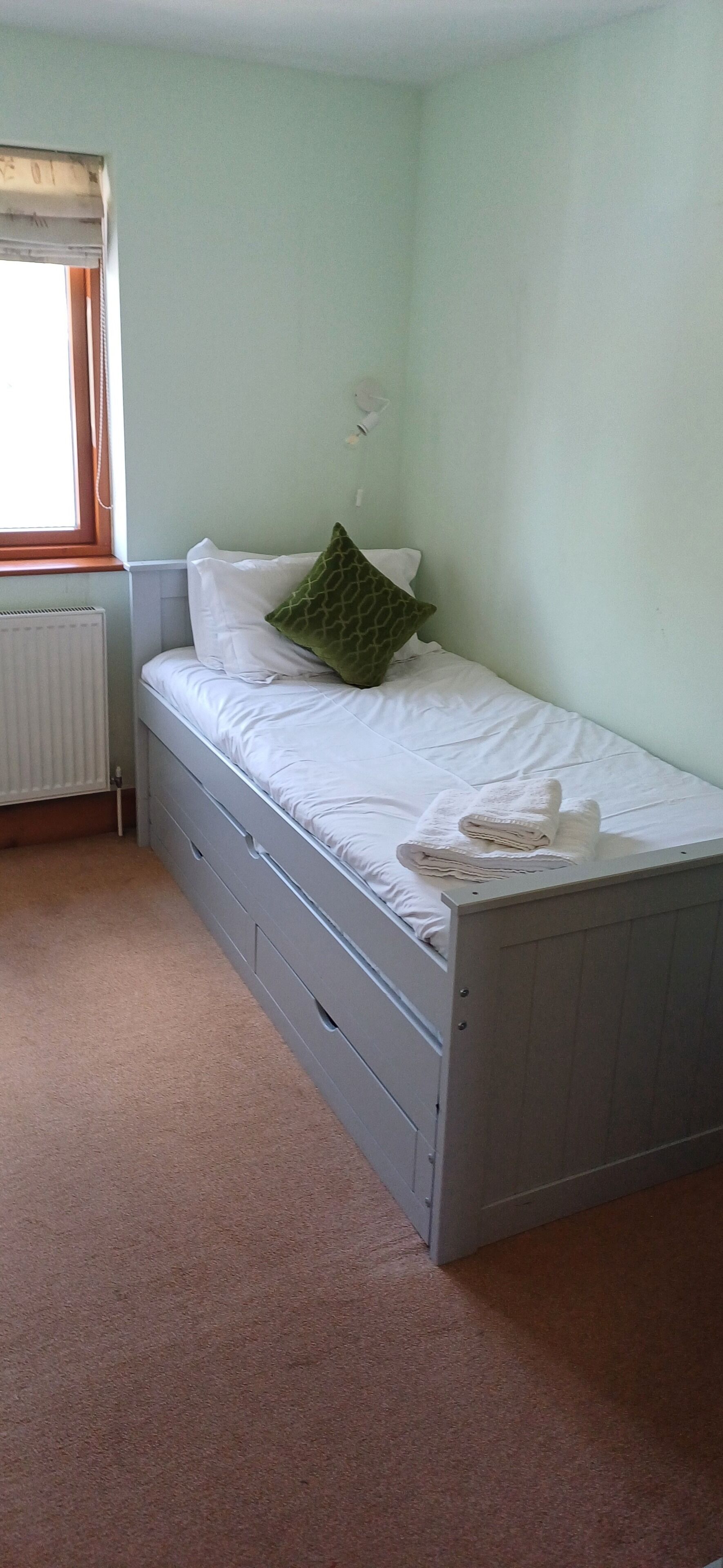 A small, single-bed room with a radiator, a window with a wooden frame, and a green pillow on the bed.