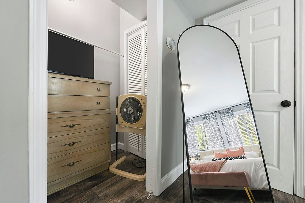 A bedroom with a bed, a dresser, a fan, and a mirror reflecting a window view.