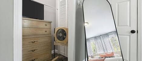 A bedroom with a bed, a dresser, a fan, and a mirror reflecting a window view.