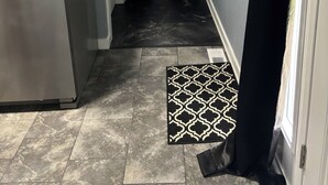 Um corredor estreito com uma geladeira à esquerda, um tapete estampado no chão e uma porta com cortina preta à direita.