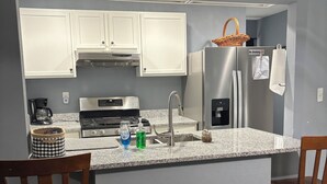 Uma cozinha compacta com armários brancos, geladeira de aço inoxidável, cafeteira e micro-ondas. Há duas cadeiras de madeira e uma pequena ilha com banquetas.