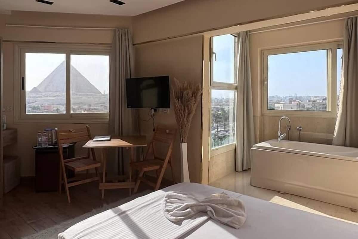 A hotel room with a bed, a desk with a chair, a TV, and two windows offering views of a pyramid and a cityscape.