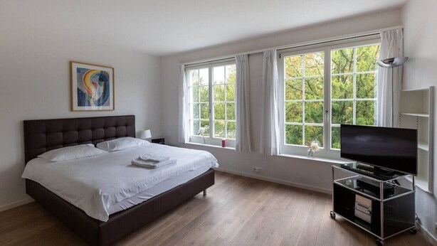 Un dormitorio con una cama grande, un televisor sobre un mueble bajo y una ventana con cortinas.