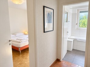 Un dormitorio con cama, un baño con lavabo y espejo, y un pasillo que conduce a una puerta.