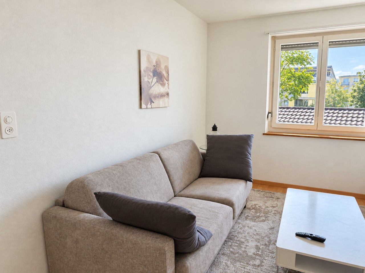 A living room with a beige sofa, two pillows, a small white table, a remote control, and a window with a view of buildings and trees.