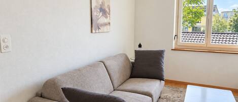 A living room with a beige sofa, two pillows, a small white table, a remote control, and a window with a view of buildings and trees.