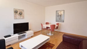 A modern living room with a flat-screen TV, a white coffee table, a red couch, and a dining area with a white table and red chairs.