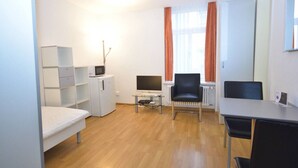 A compact room with a bed, a desk with a computer, a chair, a small refrigerator, and a radiator. - Practical studio at trendy location - Stork 22 (Zürich)