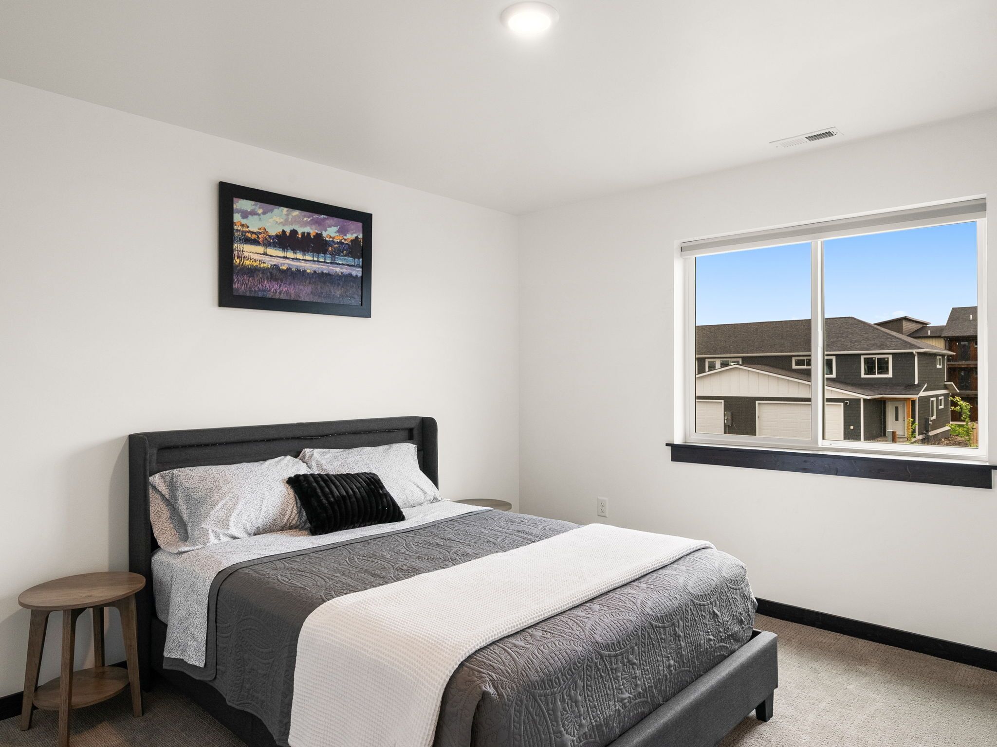 A bedroom with a bed, a nightstand, a window with a view of houses, and a framed picture on the wall.