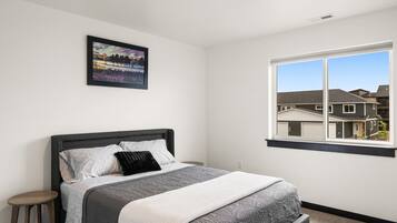 A bedroom with a bed, a nightstand, a window with a view of houses, and a framed picture on the wall.