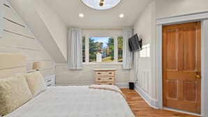 A bedroom with a large bed, a wooden dresser, a television, and a window with curtains. - Raspberry Retreat Cottage (Lovettsville)
