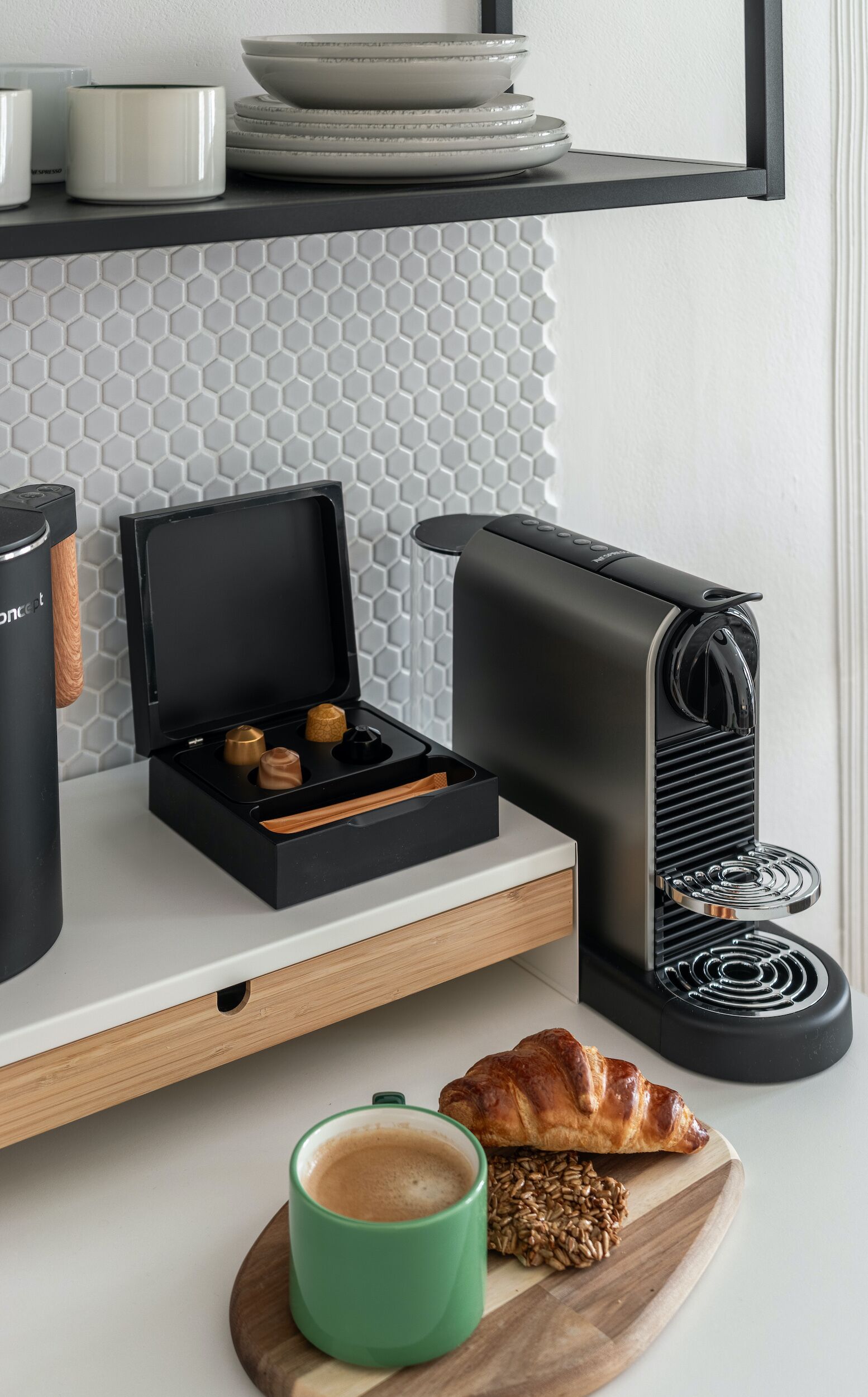 A kitchen counter with a coffee machine, a pastry, a green cup, and a tray with coffee pods.