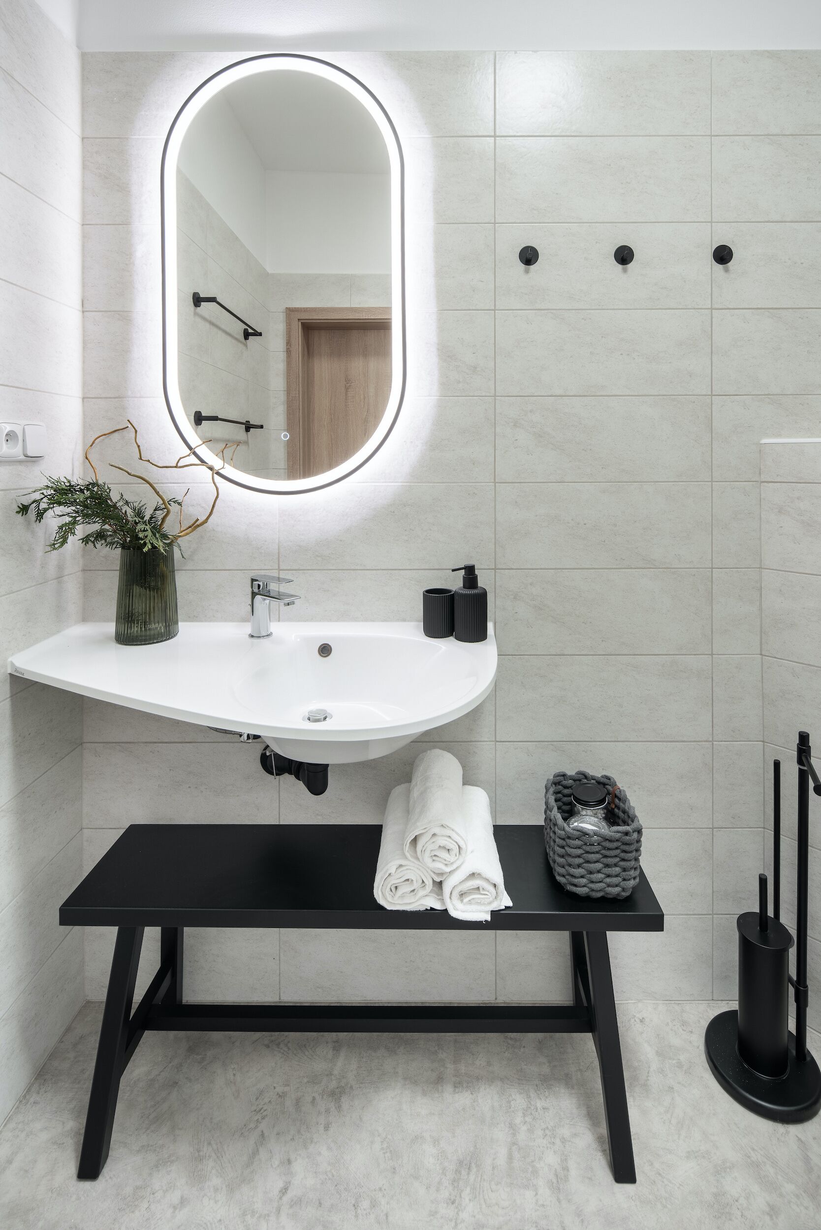 A modern bathroom with a white sink, a mirror with integrated lighting, a black bench, and a towel rack.