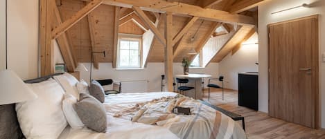 A bedroom with a high ceiling, wooden beams, a bed with a blanket and pillows, a desk, a chair, a small table, and a window.