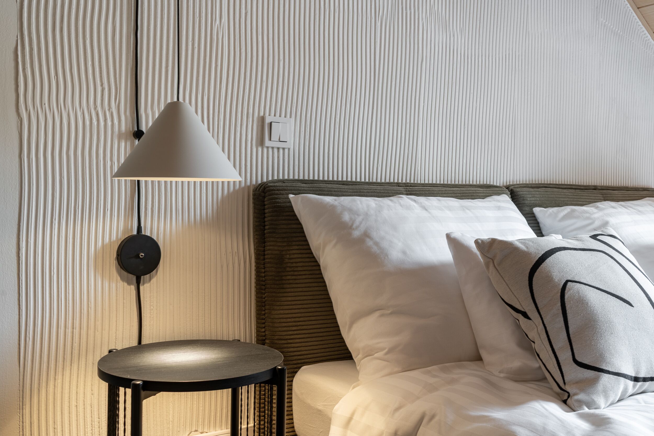 A neatly made bed with white linens and a pillow with a geometric design, a round black side table, and a wall-mounted lamp.