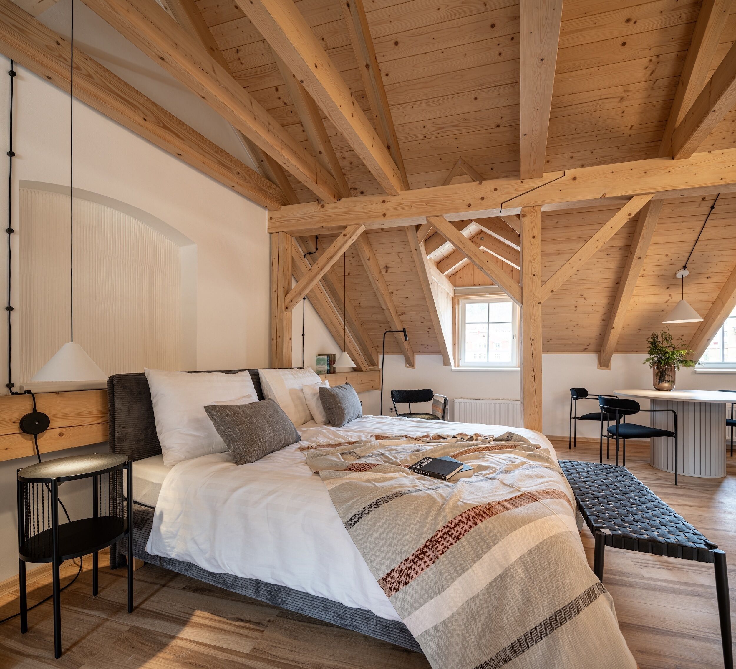 A bedroom with a wooden ceiling, a bed with a striped blanket, a bedside table, a chair, and a desk with a chair.