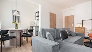 Una sala moderna con un sofá gris, sillas negras, una mesa redonda y una zona de cocina con una lámpara colgante.