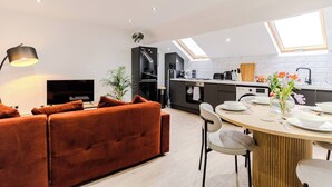 A modern living room with a sofa, a TV, and a dining area with a table set for two. - Stunning Sky view 2 BR Flat - Free WIFI, Parking (Harehills)