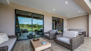 A modern living room with wicker furniture, a glass door leading to a patio, and a view of a golf course. - Disney Vacation Rental | 3BR Luxury Townhouse (Kissimmee)