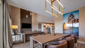 A modern dining area with a wooden table and leather chairs, a kitchen in the background, and a large painting of a historical building. - Disney Vacation Rental | 3BR Luxury Townhouse (Kissimmee)