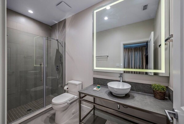 A modern bathroom with a glass-enclosed shower, a toilet, a sink with a marble countertop, and a large mirror with LED lighting.