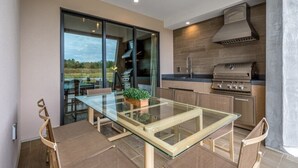 Une cuisine moderne avec une table à plateau en verre, des chaises en bois, un barbecue intégré et une porte-fenêtre coulissante donnant sur un balcon.