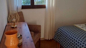 Une chambre avec un bureau en bois, une chaise en osier, un lit avec une literie à motifs et une vue sur la mer par la fenêtre.