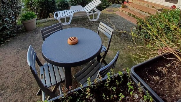 Une terrasse avec une table ronde et des chaises, un pot de fleurs et une vue sur la mer et les montagnes.