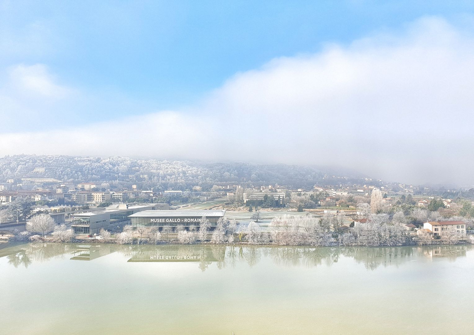 Una vista panorámica de un paisaje urbano con un edificio grande identificado como 'MUSEE GALLO - ROMAIN' reflejado en un cuerpo de agua tranquilo.
