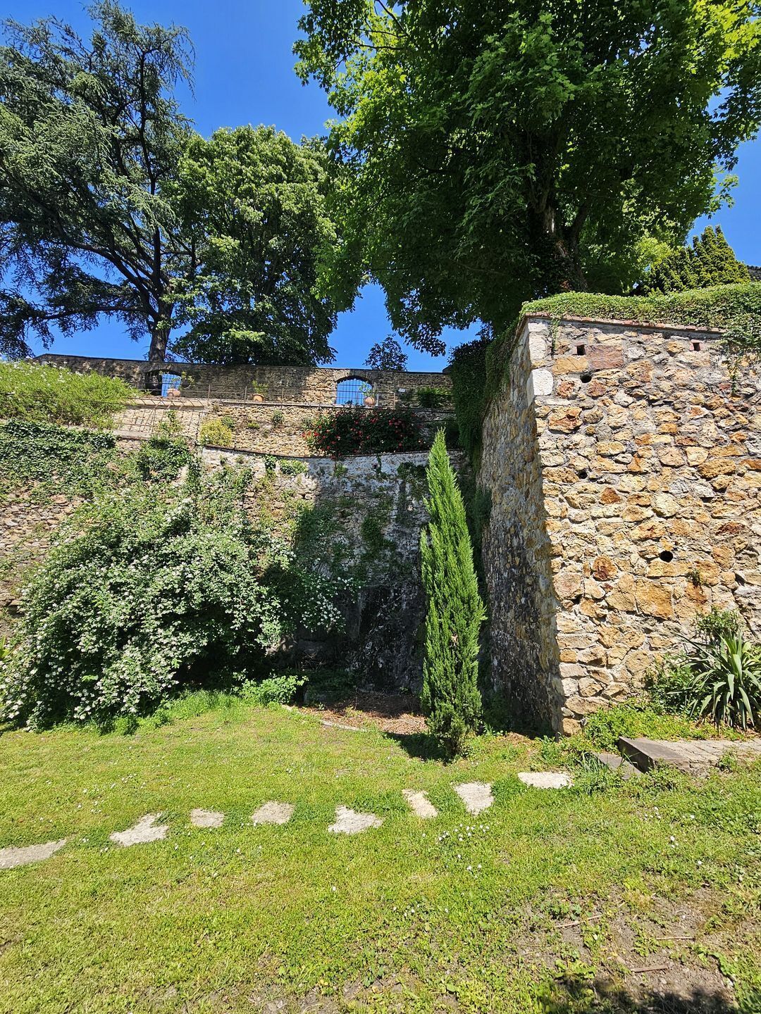 Eine Steinmauer mit einem kleinen Fenster, umgeben von Grünpflanzen und Bäumen.