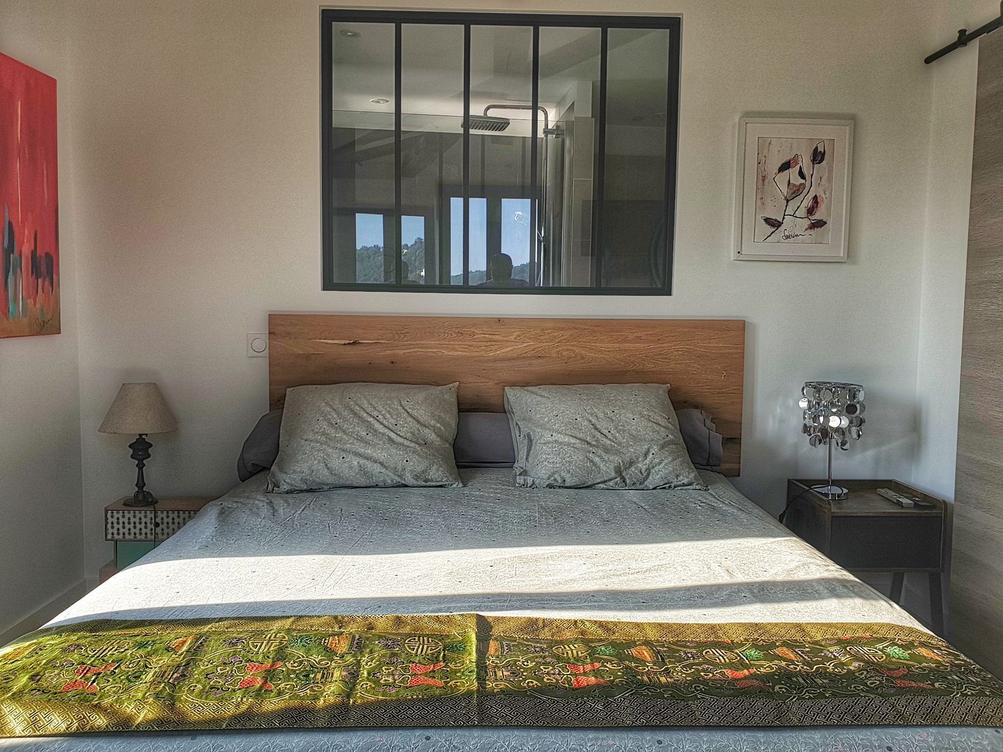 Une chambre à coucher avec une tête de lit en bois, deux oreillers, une couvre-lit à motifs, une table de chevet avec une lampe et un tableau encadré au mur.