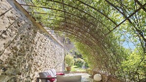 Eine Holzterrasse mit einer Bank und einem Tisch, umgeben von einer Steinmauer und überdacht durch einen Spalierbogen mit Weinlaub.