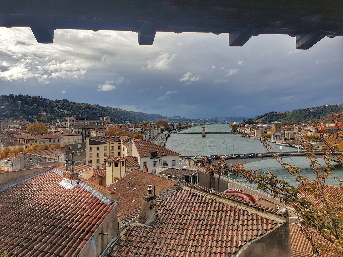 Une vue panoramique d’une ville historique, avec ses bâtiments aux toits de tuiles rouges, une rivière et un pont.