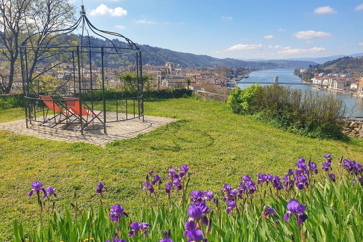 A gazebo with red chairs, surrounded by a grassy area with purple flowers, overlooking a river and a town.