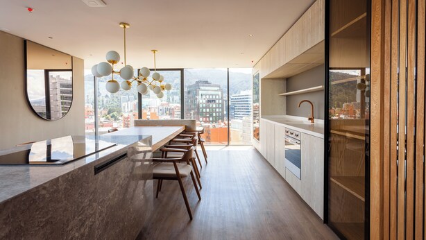 A modern kitchen with a marble countertop, wooden chairs, and a large window offering a city view.