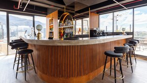 A modern bar area with a curved wooden counter, black bar stools, and a view of the city through large windows.