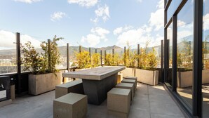 A rooftop terrace with a large concrete table and benches, surrounded by potted plants and offering a view of the city and mountains.