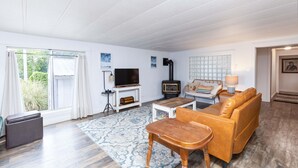 A living room with a sofa, coffee table, TV, fireplace, and a view of greenery outside. - Mermaid Crossing -Dogs OK-Kayaks-Bikes- Netart Bay (Tillamook)