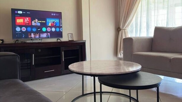 Una sala de estar moderna con un televisor de pantalla plana montado en un mueble de entretenimiento de madera oscura. Hay una mesa de centro redonda con tapa de vidrio en el centro, un sofá gris a la izquierda y un sofá beige a la derecha. La habitación tiene un ventanal con cortinas transparentes.