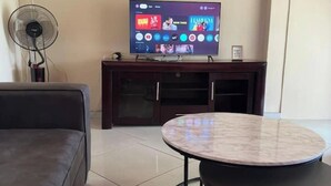 Una sala de estar con una televisión de pantalla plana sobre un mueble de madera, una mesa redonda de mármol y un sofá gris.