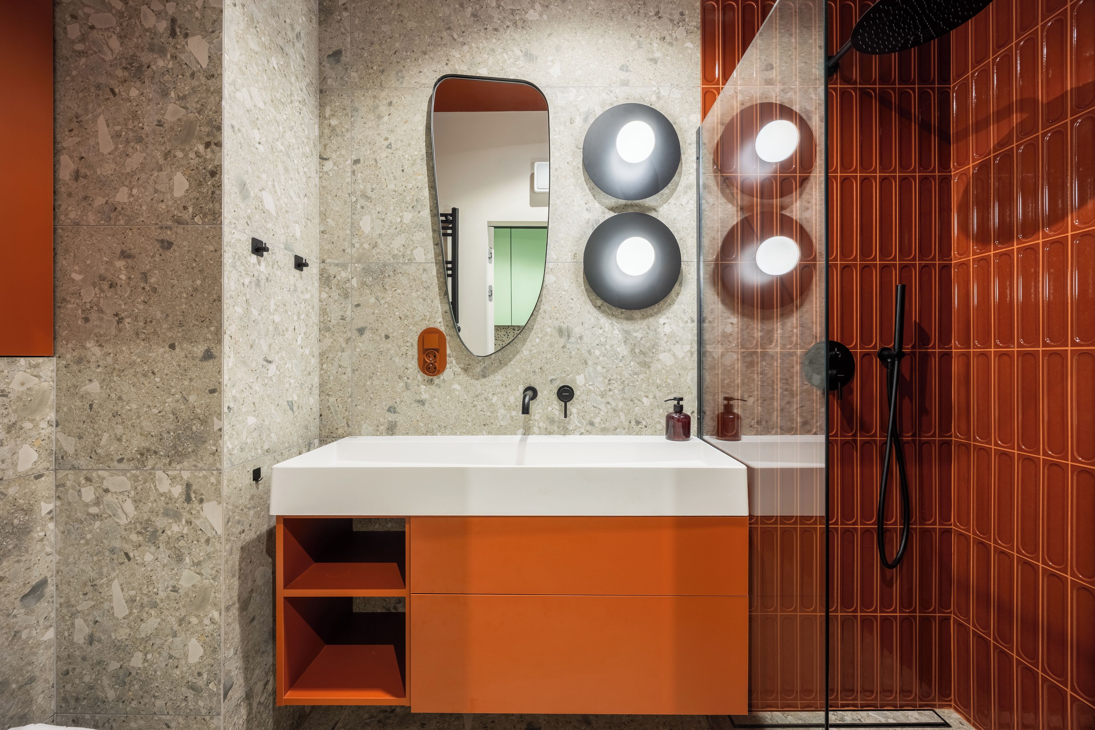 Un baño con un lavamanos blanco, un espejo, dos luces fijadas a la pared y una ducha con un revestimiento de azulejos rojos.