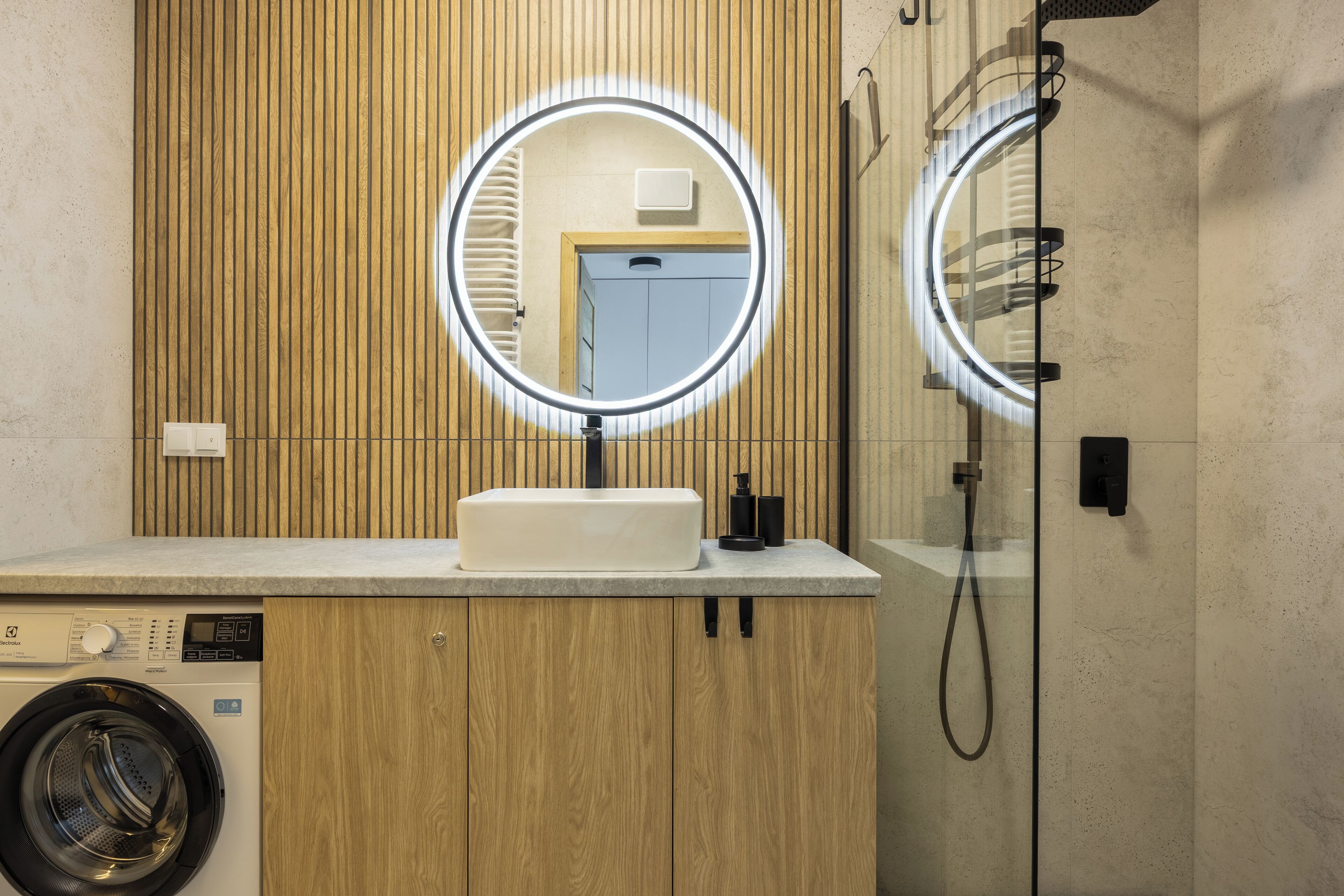 A modern bathroom with a wooden vanity, a washing machine, a sink with a mirror, and a shower area.