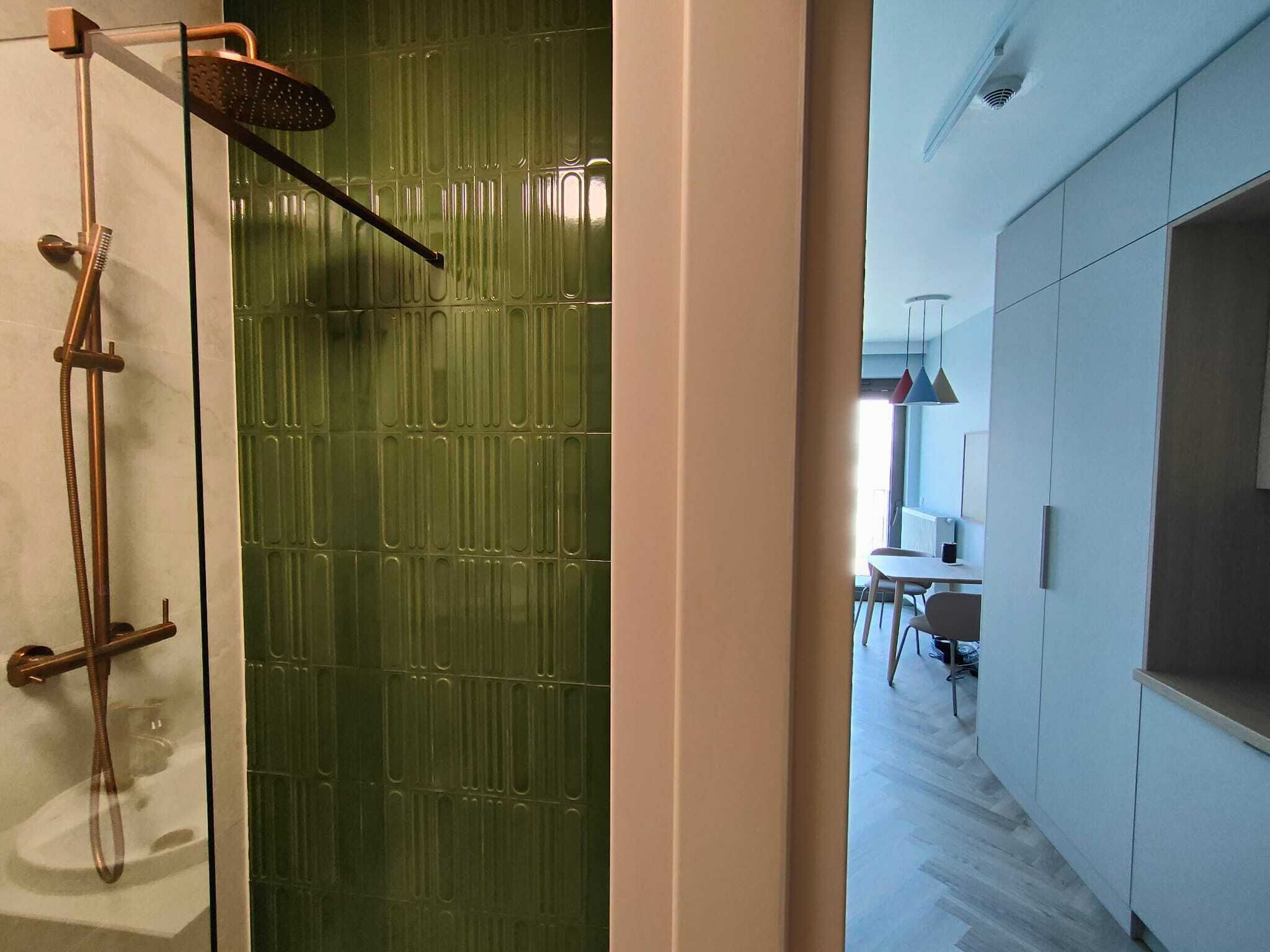 Un baño moderno con cabina de ducha revestida de azulejos verdes, un cabezal de ducha de mano y zona de ducha con mampara de cristal.