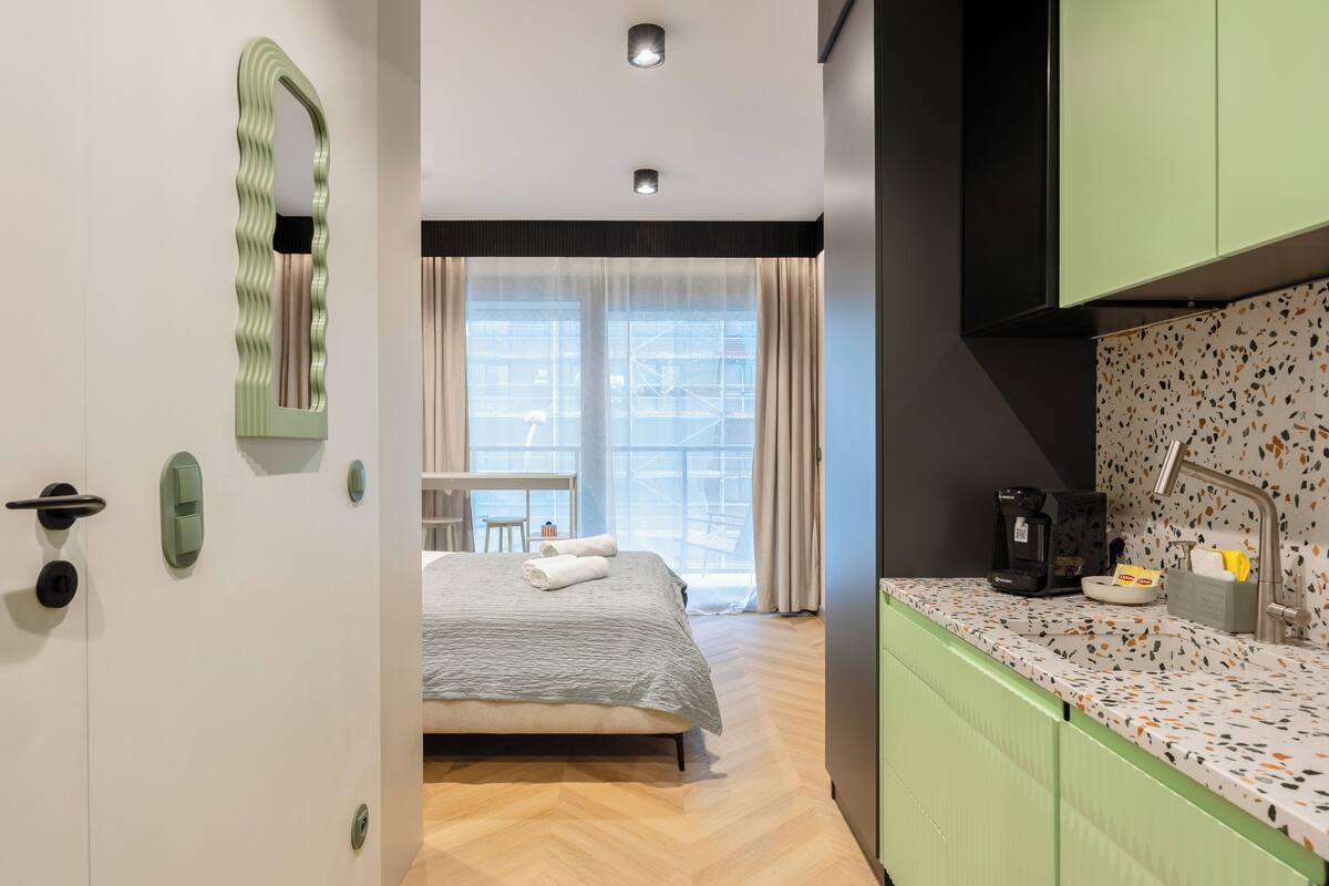 A modern hotel room with a bed, a kitchenette, and a view of the cityscape through large windows.