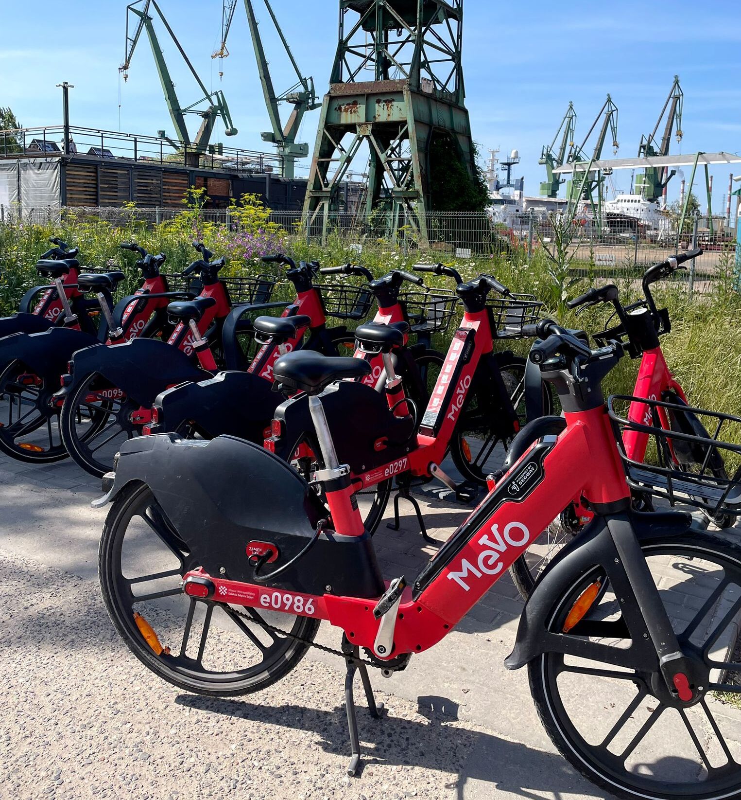Række røde elektriske cykler parkeret udendørs med en stor industriel bygning i baggrunden.