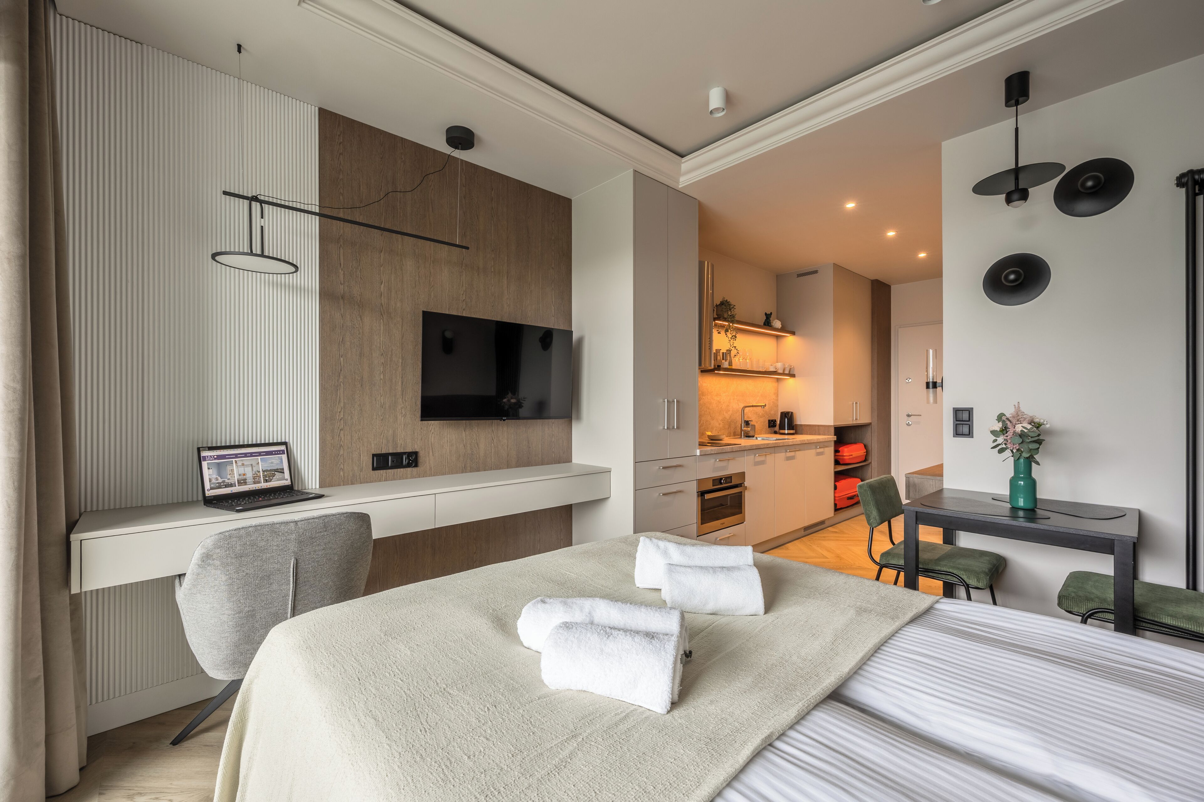 A modern hotel room with a bed, a desk with a laptop, a TV, and a kitchenette.