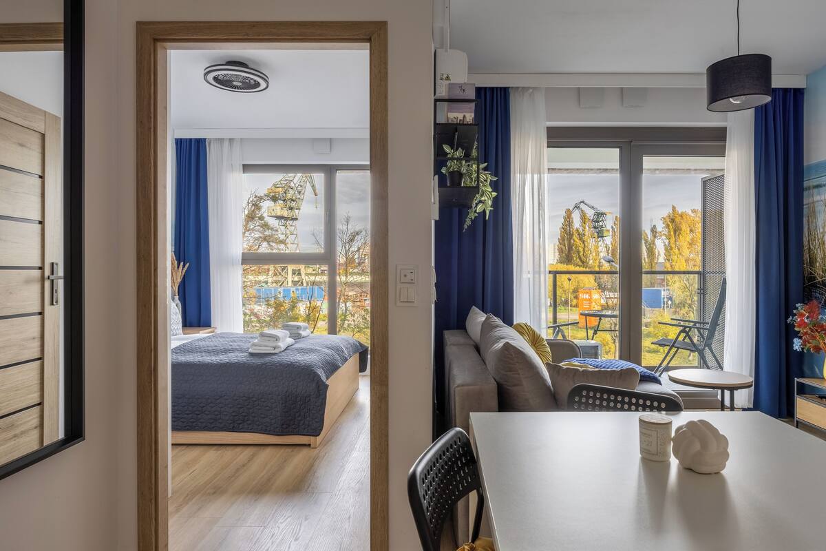 A modern hotel room with a bed, a sofa, a dining table, and a view of the outside.